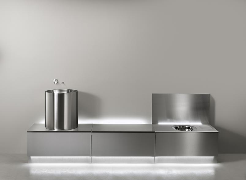 The same Abaco system in stainless steel, its flat panels closed and indistinguishable from furniture, alongside a freestanding cylindrical basin