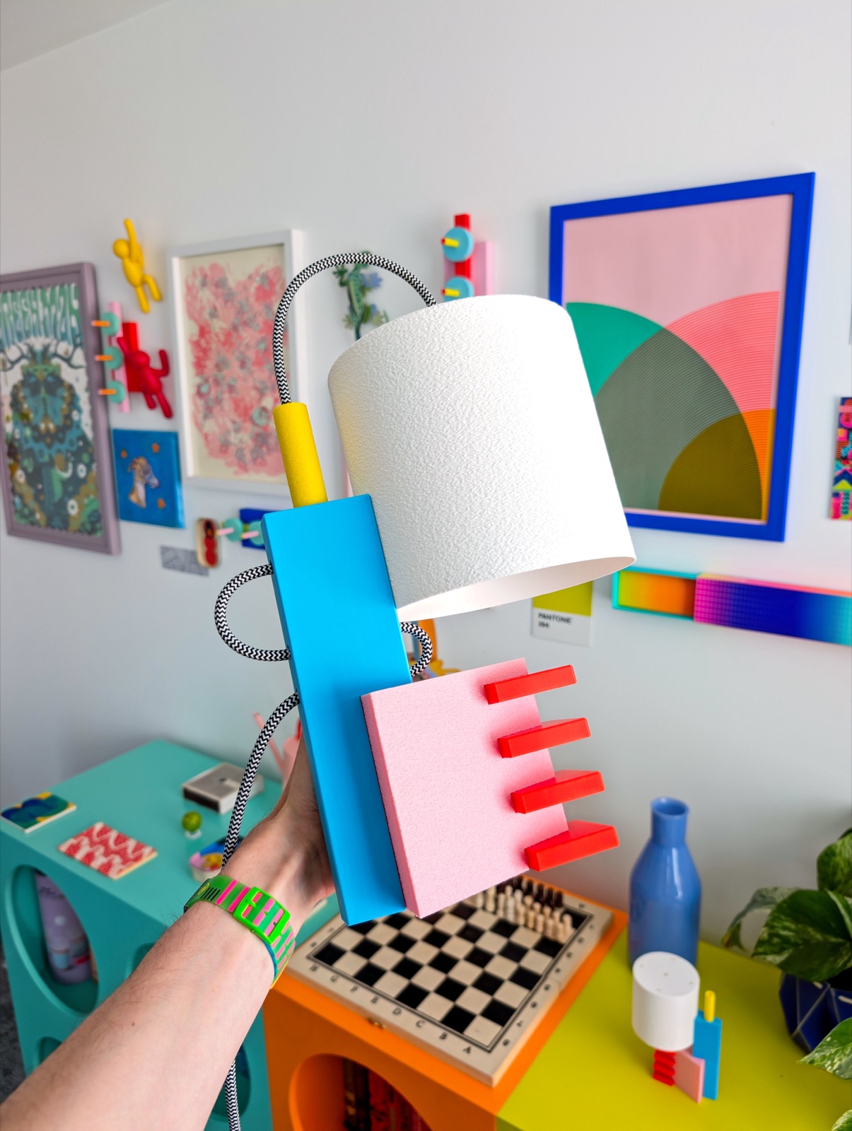 Charlie Firth's "Memphis Is Back" lamp held up against a wall of colorful Memphis-inspired art and objects, showing its blue, pink, red, and yellow geometric base