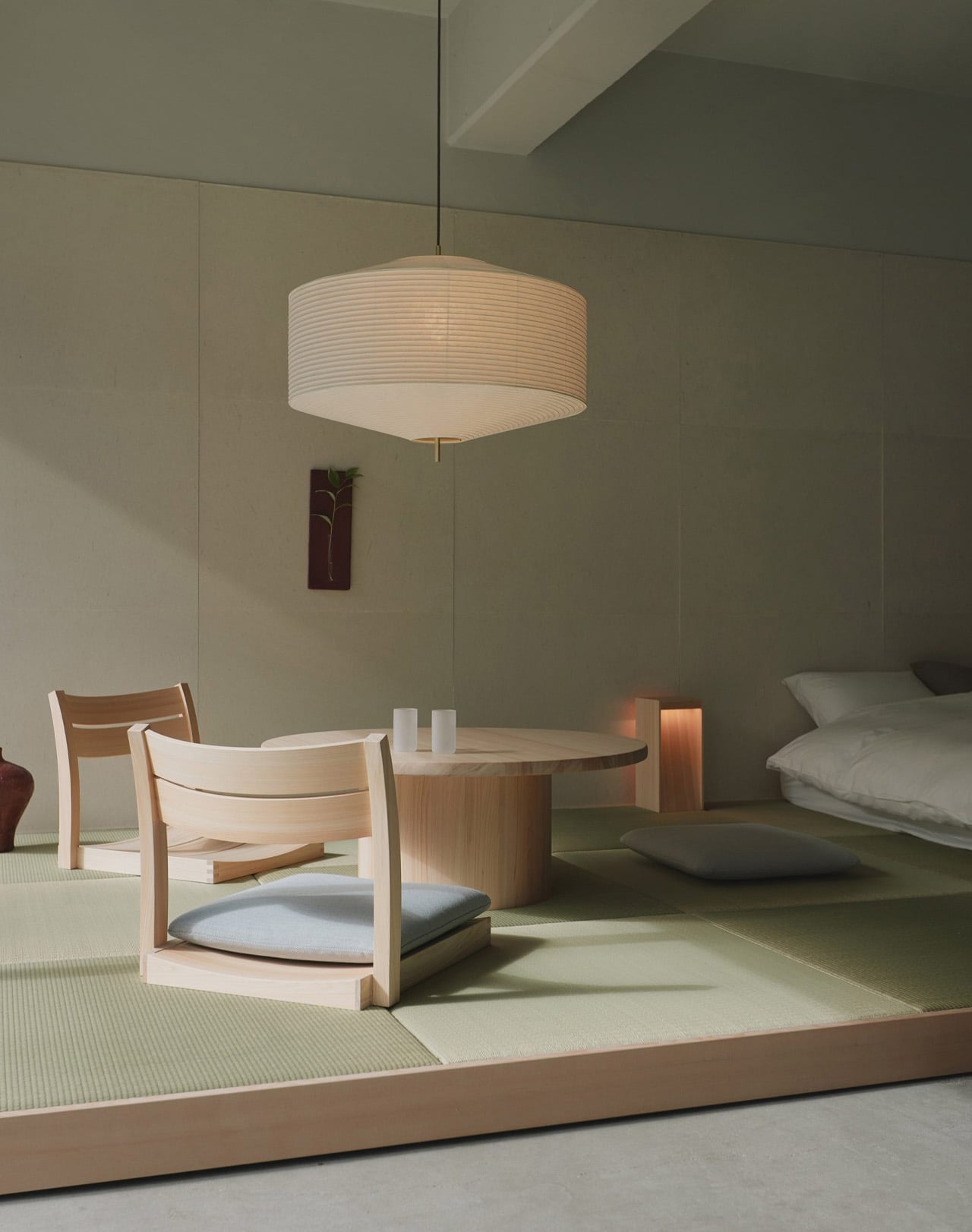 A tatami-floored room in the Oyado MAS exhibition, with two legless wooden chairs pulled up to a low round table under a paper pendant lamp, and a futon bed in the background
