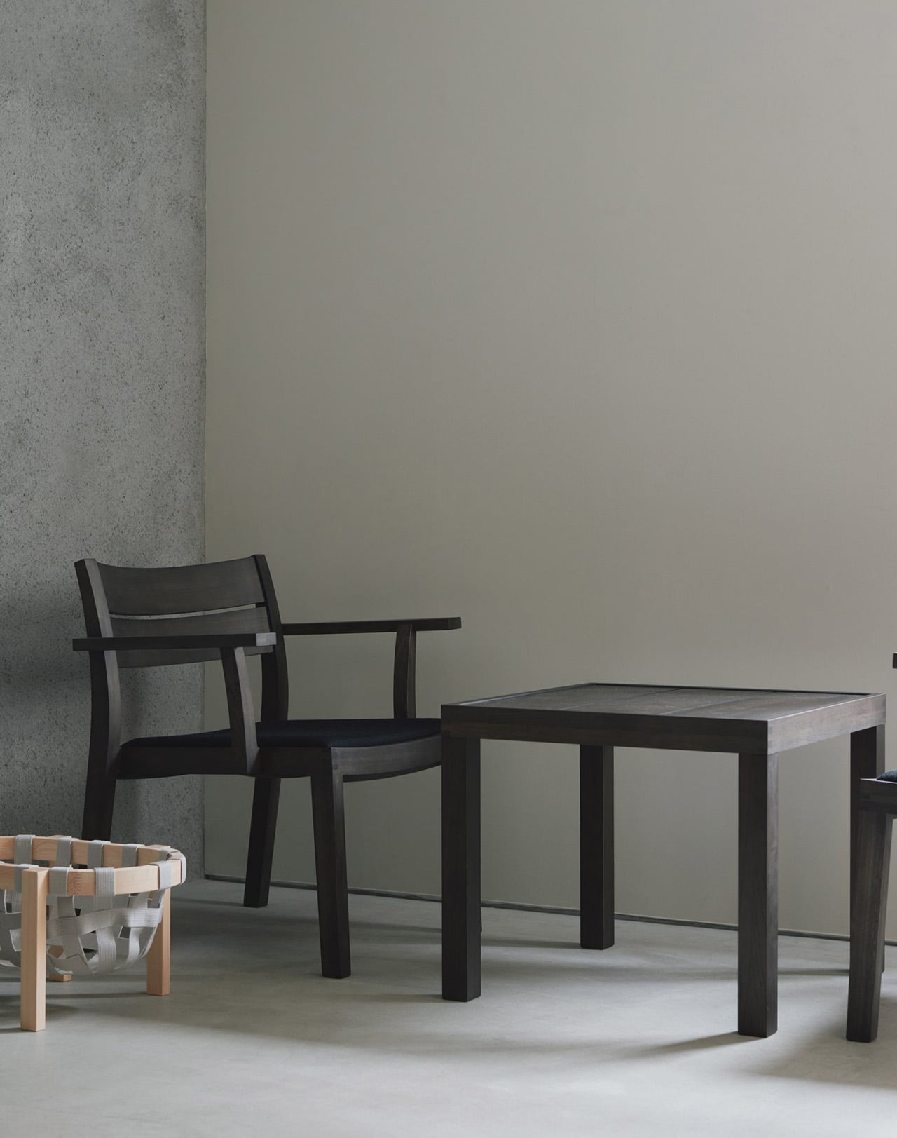 A dark-stained MAS armchair beside a matching square side table against a bare concrete wall, with a woven-strap ottoman in the corner