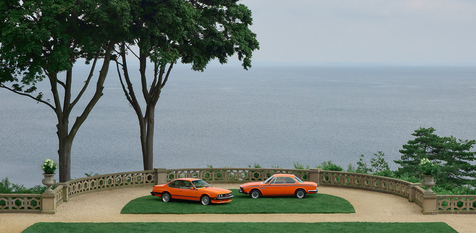 The restored 1972 BMW 3.0 CSi and 1987 BMW M6, both in Inka Orange, parked side by side overlooking the water