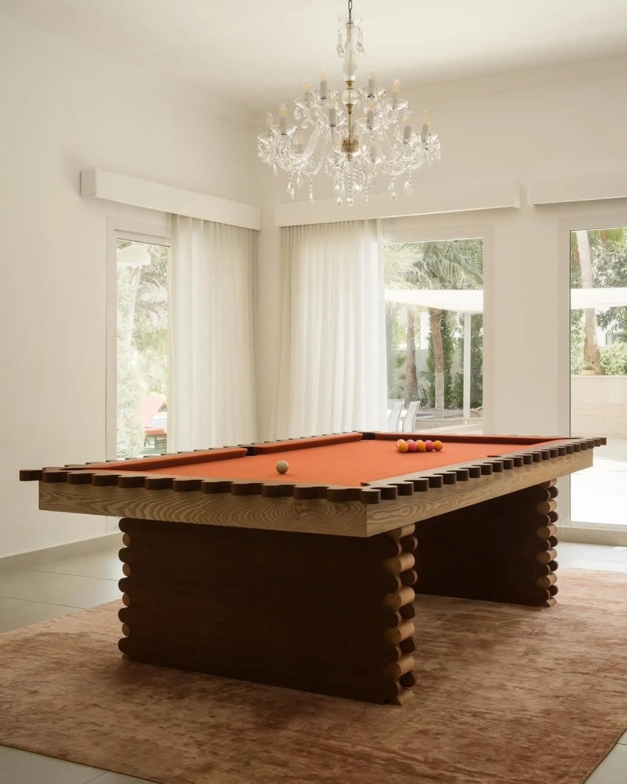 The table converted into its pool configuration, with rust-orange felt, a wooden ball tray and a triangle of balls, under a crystal chandelier