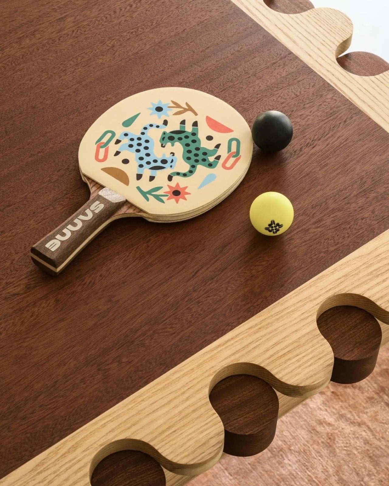 A hand-painted ping pong paddle and two balls resting on the table's dark wood surface near the fold-out net