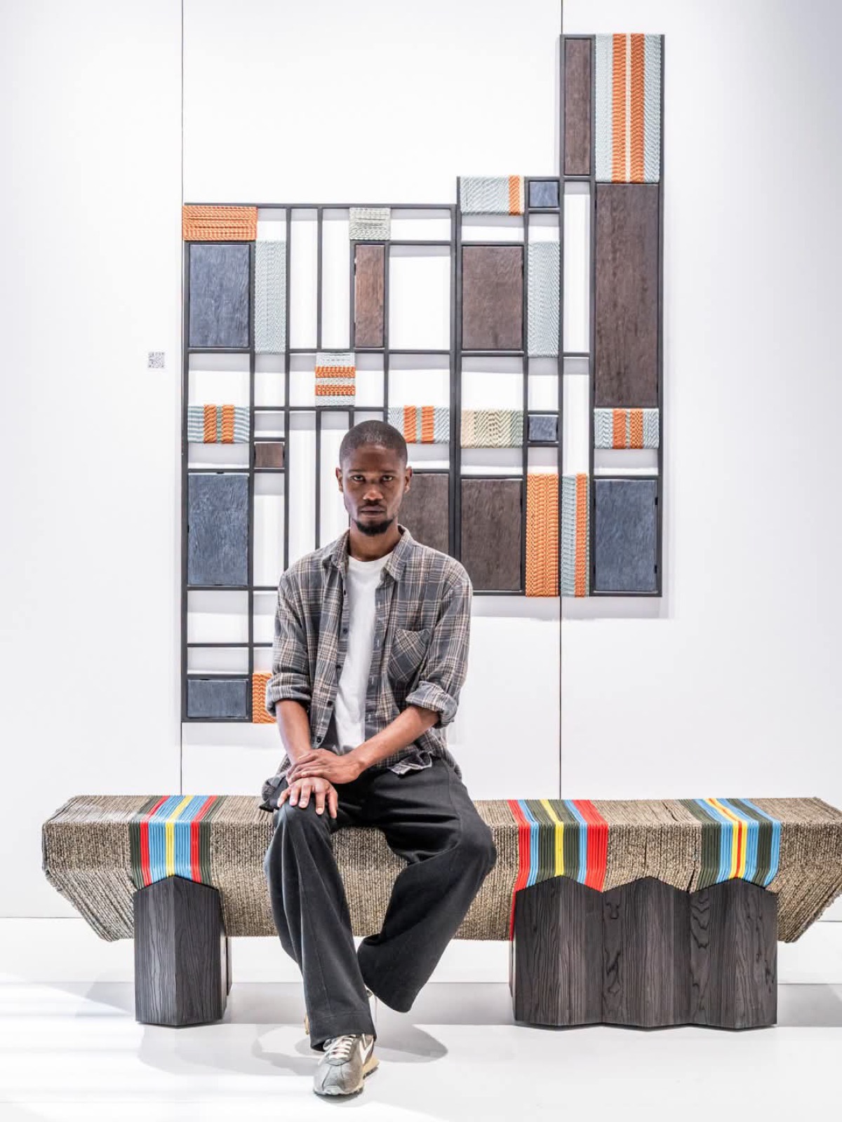 Simphiwe Mlambo seated on a striped bench from his collection, in front of a wall piece referencing his grandmother's grass mats